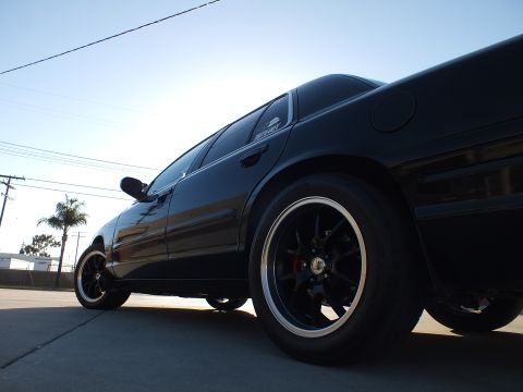 2003 Ford Crown Victoria - Close low side view of a black sedan's rear black wheel, red brake caliper and glossy bodywork in sunlight.