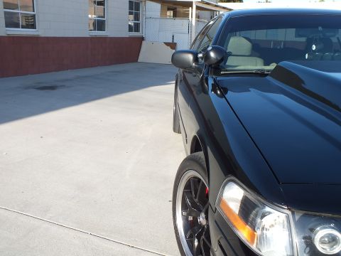 2003 Ford Crown Victoria - Close front-quarter detail of a black sedan showing its hood, side mirror, headlight and black multi-spoke wheel.