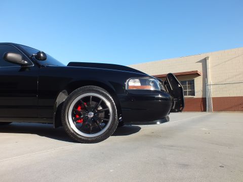 2003 Ford Crown Victoria - Low front-quarter view of a black sedan with black wheels, red caliper and push bar parked by a concrete-block wall.