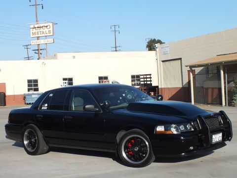 2003 Ford Crown Victoria - blacked-out full-size sedan with billet grille and push bar parked in front of an ASCO Metals industrial building.