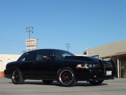 2003 Ford Crown Victoria - Front three-quarter view of a blacked-out sedan with billet grille and push bar in a lot by an ASCO Metals sign.