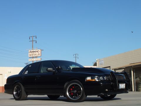 2003 Ford Crown Victoria - Side-front view of a lowered blacked-out sedan with billet grille, push bar and dark wheels near an ASCO Metals sign.