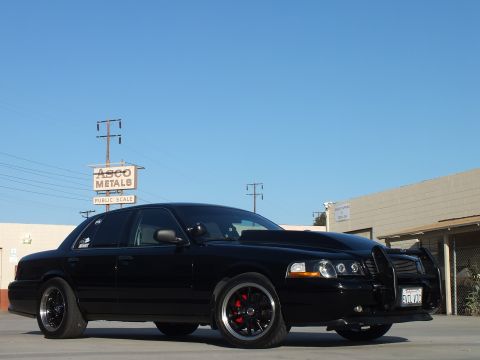 2003 Ford Crown Victoria - Front three-quarter view of a blacked-out full-size sedan with push bar and dark wheels under a clear blue sky.