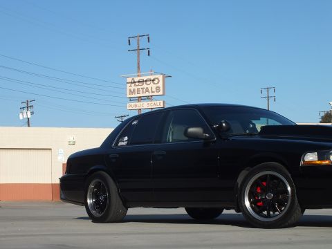 2003 Ford Crown Victoria - Side view of a lowered blacked-out sedan with dark multi-spoke wheels parked beneath an ASCO Metals public-scale sign.