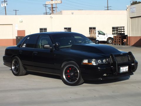 2003 Ford Crown Victoria - Front three-quarter view of a blacked-out sedan with push bar, halo headlights and a California EVIL VIC plate in an industrial lot.