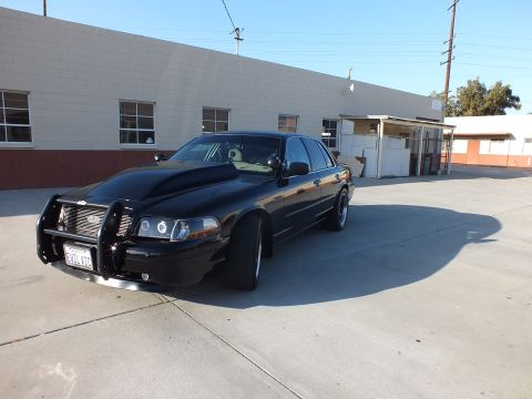 2003 Ford Crown Victoria - black Ford Crown Victoria sedan fitted with a police-style push bar and dark wheels sits on pavement beside a beige building.