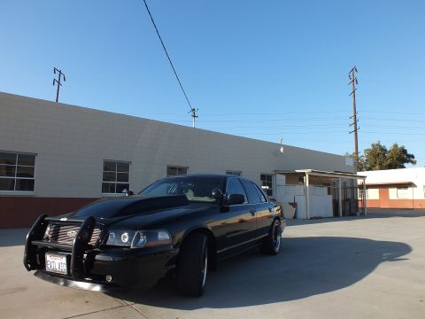 2003 Ford Crown Victoria - Front three-quarter view of a black Ford Crown Victoria with a heavy push bar, parked in an open lot under clear blue sky.