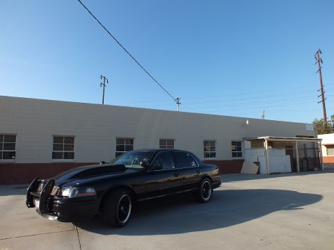 2003 Ford Crown Victoria - black Ford Crown Victoria with front push bar parked beside a long beige industrial building under a clear sky.