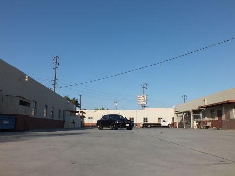 2003 Ford Crown Victoria - black sedan parked in an industrial yard near a white flatbed truck and a tall lit sign under blue sky.