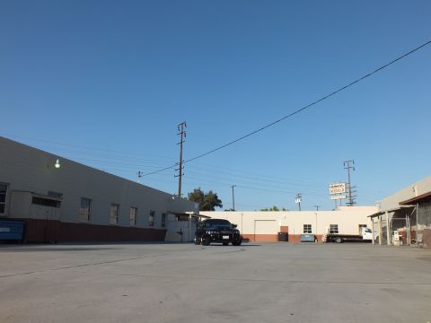 2003 Ford Crown Victoria - small black sedan sits far across a broad concrete lot flanked by warehouse buildings and utility poles under clear sky.