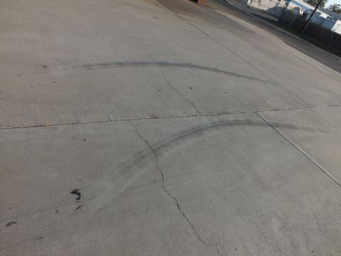 2003 Ford Crown Victoria - Two large curved tire skid marks arc across a wide gray concrete lot bordered by a fence in the distance.