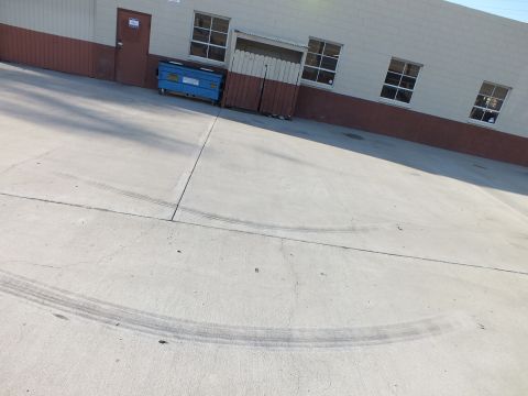 2003 Ford Crown Victoria - Curved tire skid marks streak across a concrete lot in front of a warehouse wall and a blue dumpster.