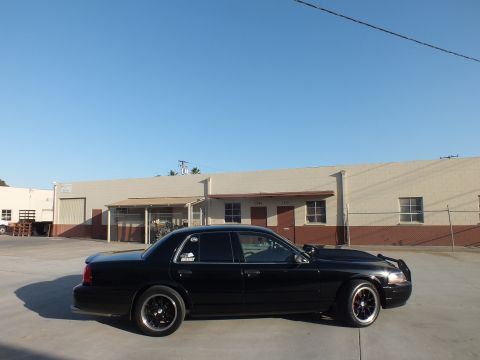 2003 Ford Crown Victoria - lowered black full-size sedan on black wheels sits in side profile in a sunlit industrial lot near warehouses.