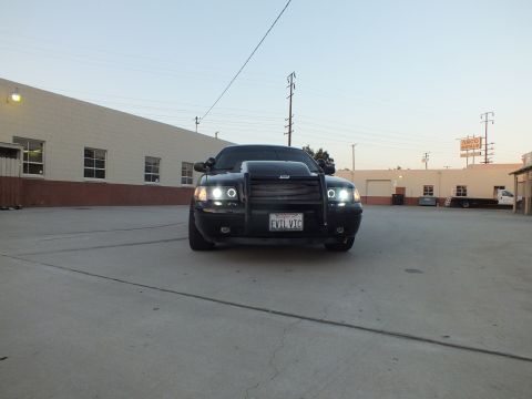2003 Ford Crown Victoria - black Ford Crown Victoria with push bar and halo headlights sits in an industrial lot near an Asco Metals sign at dusk.