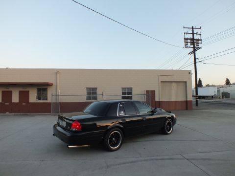 2003 Ford Crown Victoria - Rear three-quarter view of a black full-size sedan on black wheels parked beside a tan industrial building.