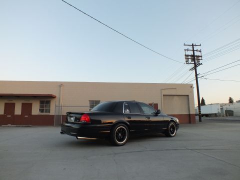 2003 Ford Crown Victoria - Rear three-quarter view of a black full-size sedan on dark wheels parked beside a tan building.