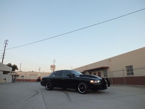 2003 Ford Crown Victoria - Front three-quarter view of a black full-size sedan with headlights on, parked in an industrial lot.