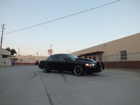2003 Ford Crown Victoria - Front three-quarter view of a black full-size sedan with headlights illuminated parked near a tan building.