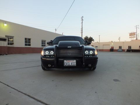 2003 Ford Crown Victoria - Head-on view of a black sedan with a push bar, halo headlights and an EVIL VIC plate parked in a wide lot.