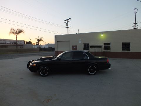 2003 Ford Crown Victoria - Side profile of a black full-size sedan on dark wheels parked beside a tan industrial building at dusk.