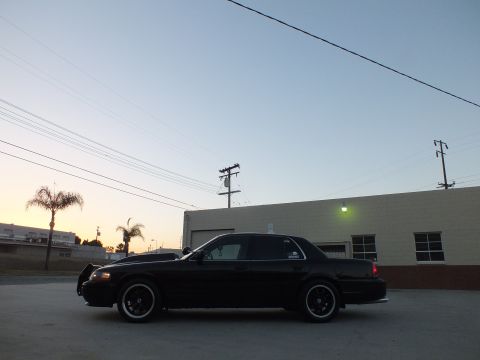 2003 Ford Crown Victoria - Driver-side profile of a black full-size sedan with headlights on, parked in a wide paved lot at dusk.