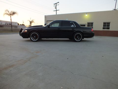 2003 Ford Crown Victoria - Side profile of a lowered black full-size sedan on black wheels parked near a building at sunset.