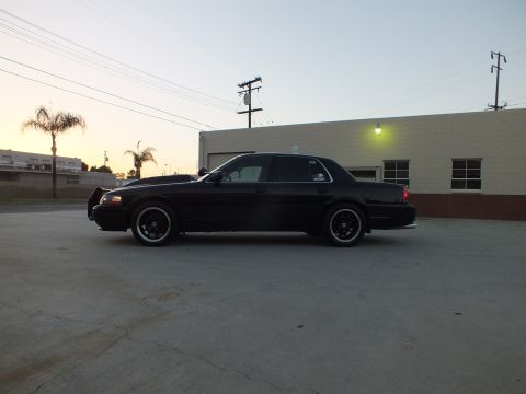 2003 Ford Crown Victoria - Driver-side profile of a black full-size sedan with headlights lit, parked beside an industrial building.