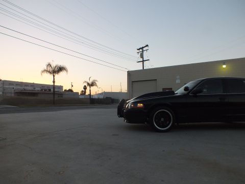 2003 Ford Crown Victoria - Front three-quarter view of a black sedan with a brush guard and headlights on, parked in a lot at sunset.