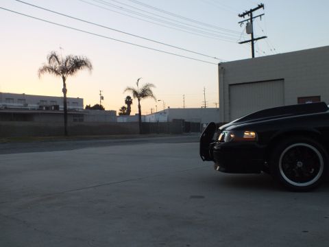 2003 Ford Crown Victoria - black sedan sits at the edge of a concrete industrial lot at dusk, with palm trees and warehouse buildings behind it.