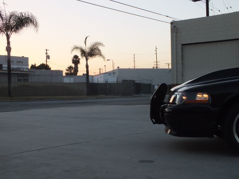 2003 Ford Crown Victoria - amber-lit front corner of a black sedan in an industrial yard at dusk, palm trees and a warehouse in the background.