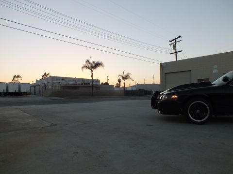 2003 Ford Crown Victoria - black sedan with a dark alloy wheel parked in a concrete lot, warehouses and a hazy sunset sky behind.