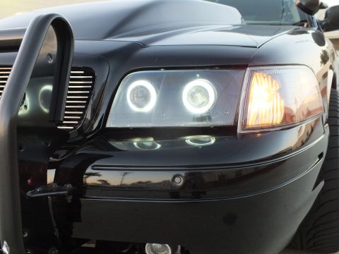 2003 Ford Crown Victoria - Detail of a black car's halo projector headlight glowing with an amber turn signal lit, framed by a black grille guard.