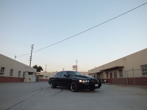 2003 Ford Crown Victoria - black sedan parked diagonally in a wide concrete industrial lot, beige warehouses and power lines under a pale dusk sky.