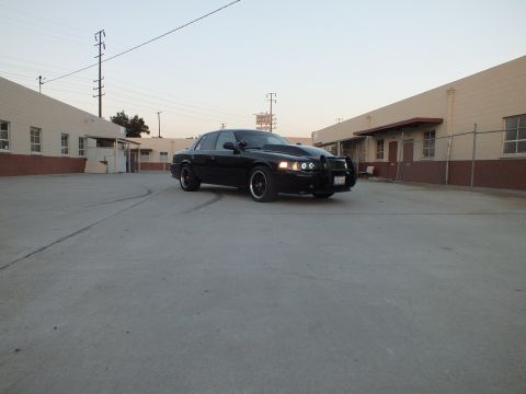 2003 Ford Crown Victoria - black sedan with headlights on sits in a concrete industrial yard flanked by beige warehouses at dusk.