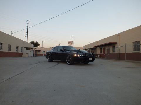 2003 Ford Crown Victoria - black sedan showing glowing halo headlights parked in a broad industrial lot between warehouses at dusk.