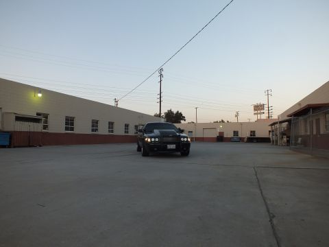 2003 Ford Crown Victoria - black sedan faces the camera with headlights lit in a large empty concrete industrial yard at dusk.