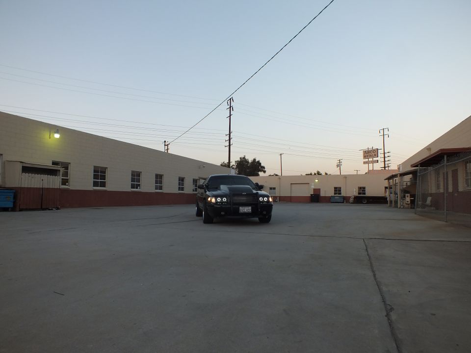Black sedan facing camera with headlights on in large industrial yard at dusk