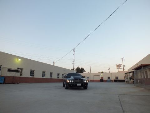 2003 Ford Crown Victoria - black sedan sits centered in a wide concrete lot beneath an overhead wire, warehouses on both sides at dusk.