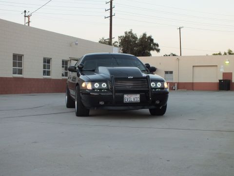 2003 Ford Crown Victoria - Front view of a black sedan with a grille guard, glowing halo lights and California plate EVIL VIC in an industrial lot.