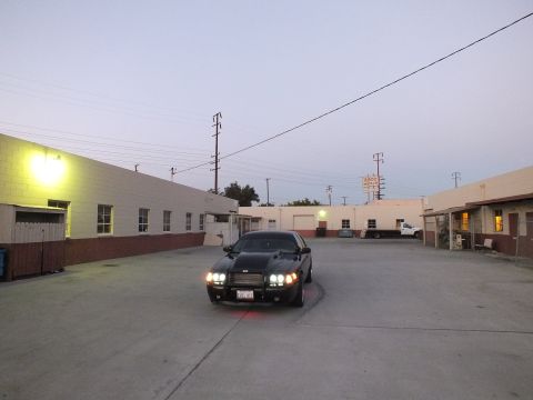 2003 Ford Crown Victoria - black sedan with headlights lit and red underglow sits in a concrete industrial yard at twilight.