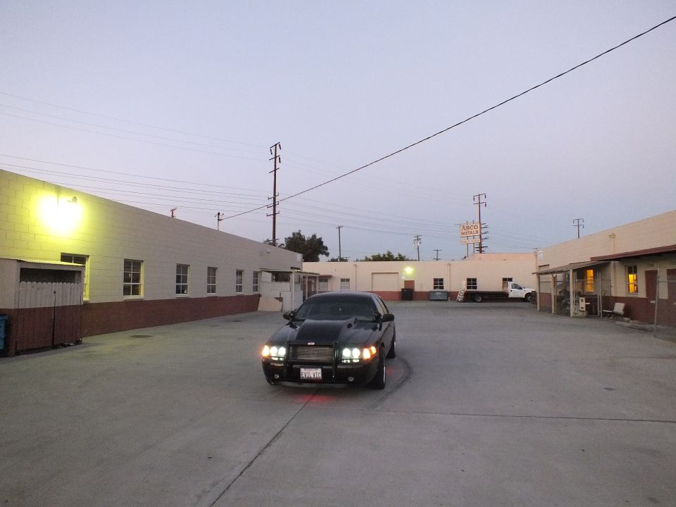 Black sedan with headlights on and red underglow in industrial lot at twilight