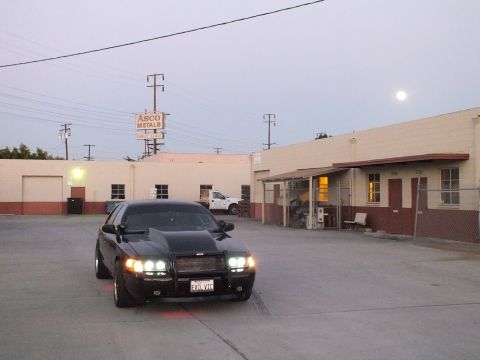 2003 Ford Crown Victoria - black sedan with headlights on parked near an ASCO Metals warehouse at dusk, the moon rising in the sky.
