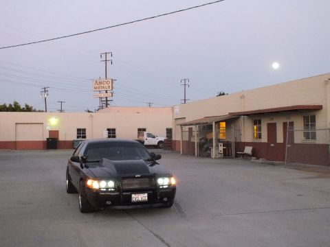 2003 Ford Crown Victoria - Front three-quarter of a black sedan with headlights lit in an industrial lot near an ASCO Metals sign, moon overhead.