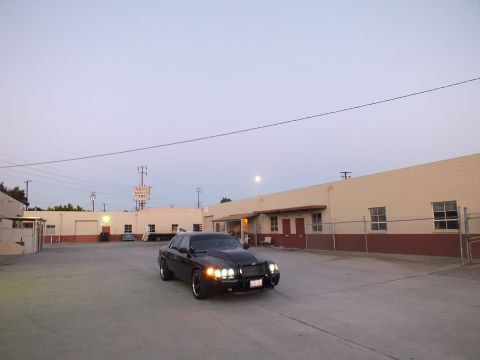 2003 Ford Crown Victoria - black sedan with headlights on sits in a wide concrete industrial lot with the moon rising over warehouses at dusk.