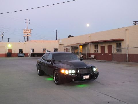 2003 Ford Crown Victoria - black sedan glows with green underglow and lit headlights in an industrial lot near an ASCO Metals sign at dusk.