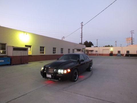 2003 Ford Crown Victoria - black sedan with lit headlights and red underglow parked in a concrete industrial lot near warehouses at dusk.