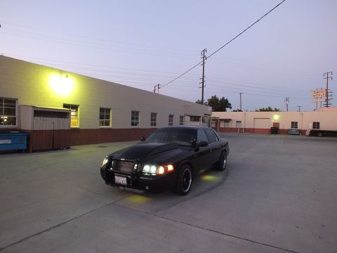 2003 Ford Crown Victoria - blacked-out full-size sedan with green underglow sits in an empty industrial parking lot under a glowing wall light at dusk.