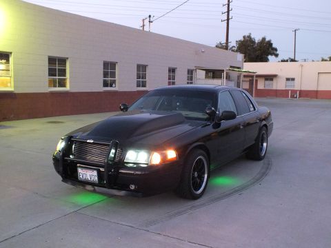 2003 Ford Crown Victoria - black full-size sedan with a chrome push-bar grille guard, green underglow and an 'EVIL VIC' license plate parked by an industrial building.
