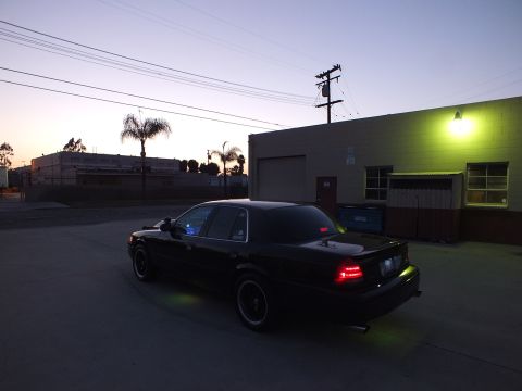 2003 Ford Crown Victoria - black full-size sedan shown from the rear three-quarter with glowing taillights, parked in an industrial lot at sunset.