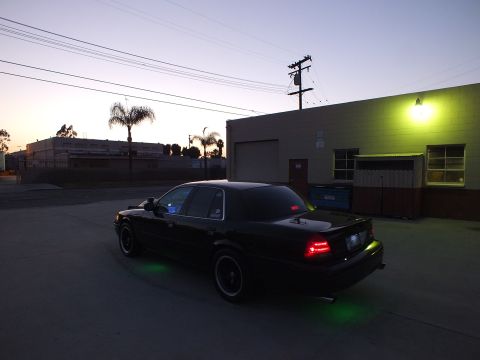 2003 Ford Crown Victoria - black full-size sedan with green underglow and lit red taillights, parked in an industrial lot at dusk.
